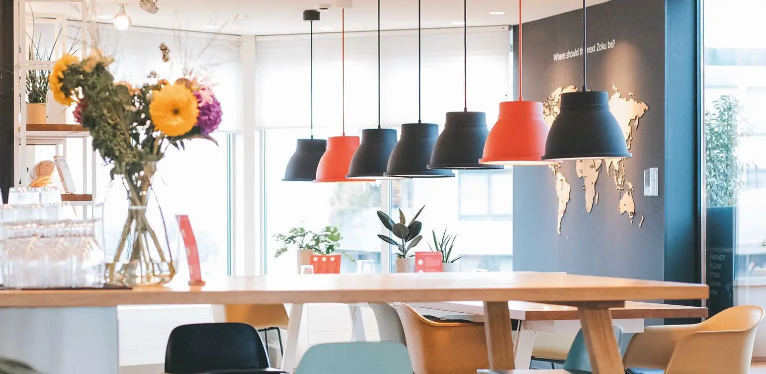 Espacio moderno con lámparas colgantes negras y rojas, mesa de madera, sillas de colores y decoración con plantas.