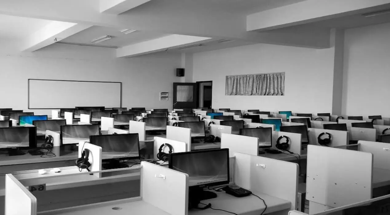 Sala de oficinas con numerosas estaciones de trabajo tipo cubículo, cada una equipada con monitor, teclado y auriculares, distribuidas en filas ordenadas dentro de un espacio amplio y bien iluminado, con paredes blancas, pizarrón y ventana cubierta por cortina.