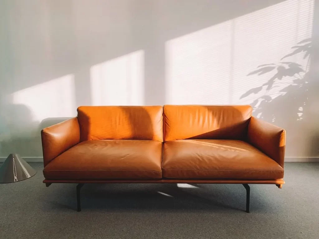 Sofá de diseño contemporáneo de cuero color marrón claro, de dos plazas, líneas rectas y patas metálicas negras, ubicado en una sala luminosa con pared blanca y luz natural proyectando sombras geométricas sobre el fondo.