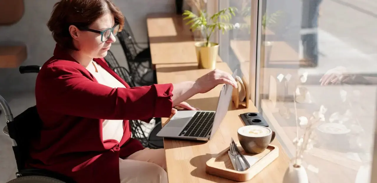 Mujer en silla de ruedas trabajando en una laptop junto a una ventana en una cafetería, con una taza de café y utensilios sobre la mesa