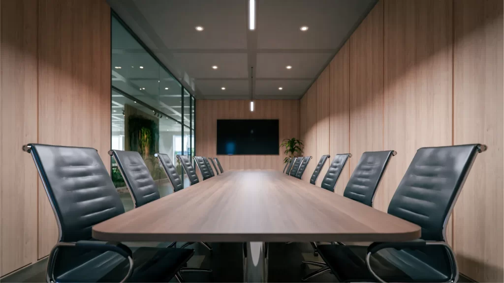 Una elegante sala de reuniones con una mesa de madera alargada, rodeada de modernas sillas negras de diseño ergonómico con estructura metálica. Las paredes están revestidas en madera, creando un ambiente cálido y sofisticado. Al fondo, un televisor está montado en la pared, y una planta añade un toque de frescura al espacio. Un panel de vidrio permite la entrada de luz natural desde otra área de la oficina. La iluminación empotrada en el techo y una lámpara lineal brindan una atmósfera equilibrada y profesional.