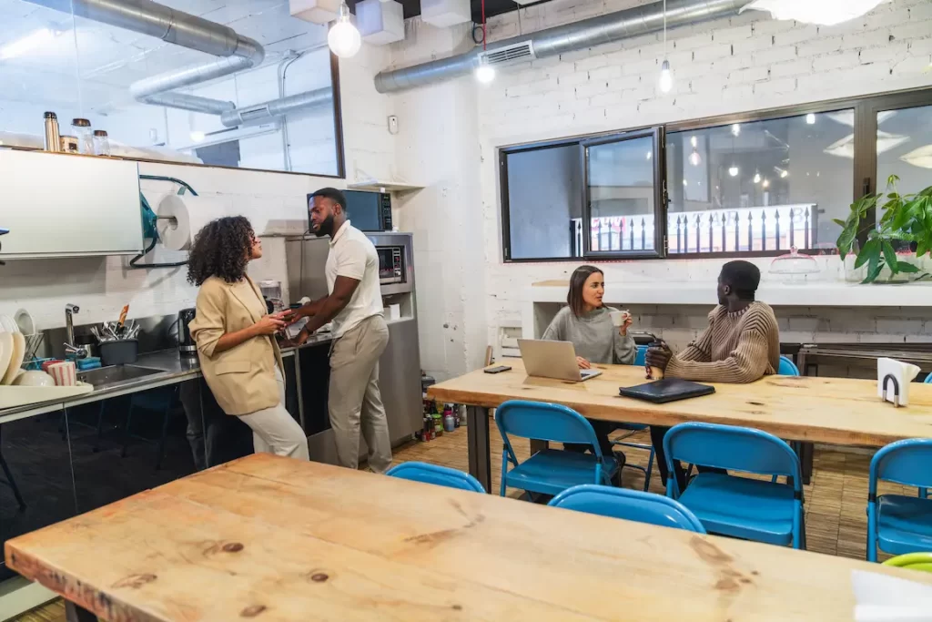 Personas conversando y trabajando en un espacio colaborativo moderno con cocina y mesas de madera.